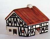 Das Bild zeigt ein detailliertes Modell einer Mhle im Fachwerkstil mit rotbraunen Dach. Die Fachwerkstruktur ist deutlich sichtbar, mit dunklen Holzbalken. Das Haus ist klein und knnte ein Sammlerstck oder Dekorationsartikel sein. Das Modell ist wahrscheinlich aus Pozellan oder einem hnlichen Material handwerklich gefertigt und wirkt sehr hochwertig. Die Farbgebung ist einfach, aber effektiv, mit dem Kontrast zwischen dem roten Dach und den hellen Wnden. Das Modell vermittelt ein Gefhl von traditioneller Architektur und handwerklichem Knnen. Es knnte ein Beispiel fr ein typisch deutsches Fachwerkhaus darstellen und Nostalgie oder ein Interesse an Architekturgeschichte hervorrufen. Die Einfachheit des Designs unterstreicht die Details der Konstruktion. Es ist wohl auch als Teelichthaus zu nutzen und von hinten zu beleuchten.