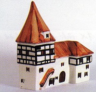 Das Bild zeigt ein detailreiches Modell eines Stadttores, das im Stil eines Fachwerkhauses gestaltet ist. Das Haus ist aus Porzellan gefertigt und wahrscheinlich ein Souvenir oder ein Sammlerstck. Das Hauptgebude ist wei verputzt mit aufflligen schwarz-weien Fachwerkwnden, die typisch fr die Architektur bestimmter Regionen Deutschlands sind. Das Dach ist rotbraun und spitz. Ein kleiner Turm mit hnlicher Dachgestaltung ist mit dem Hauptgebude verbunden. Vor dem Haus ist eine kleine Rampe oder Treppe dargestellt, die mglicherweise auf einen Zugang zum Gebude hinweist. Die Farbgebung ist einfach, aber wirkungsvoll und betont die traditionellen Elemente des Gebudes. Das Modell wirkt handwerklich gefertigt und detailreich, die einzelnen Elemente sind sorgfltig ausgearbeitet. Es ist ein charmantes Beispiel fr miniaturisierte Architektur, das mglicherweise eine spezifische historische Gebudeform reprsentiert oder einfach eine stilisierte Darstellung eines traditionellen deutschen Fachwerkhauses ist.  Es ist wohl auch als Teelichthaus zu nutzen und von hinten zu beleuchten.
